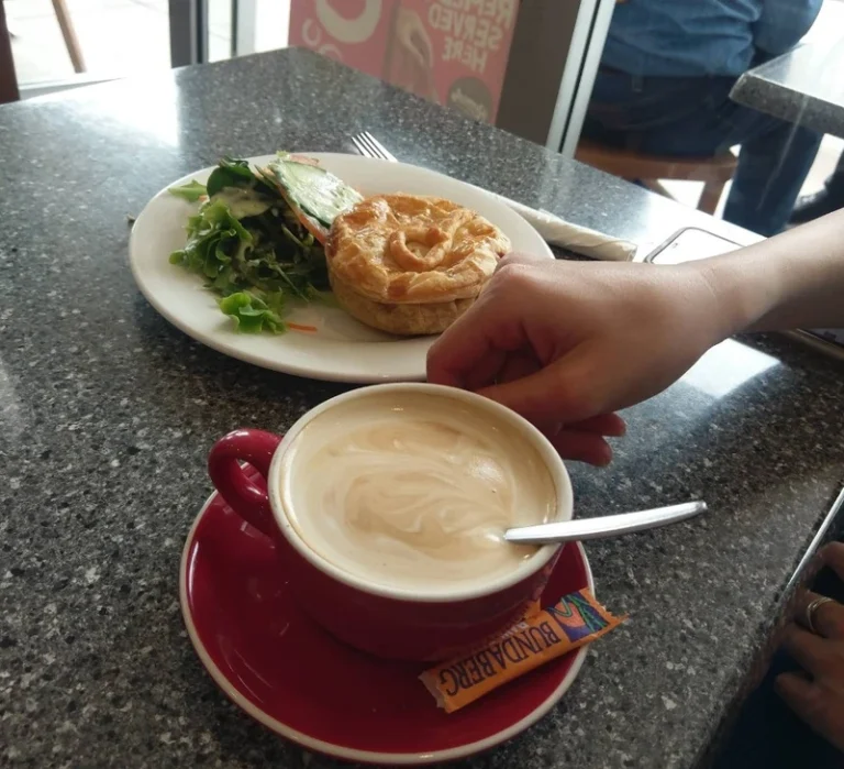 Hand holding a creamy cappuccino in a red cup with a savory meat pie and fresh salad on a plate